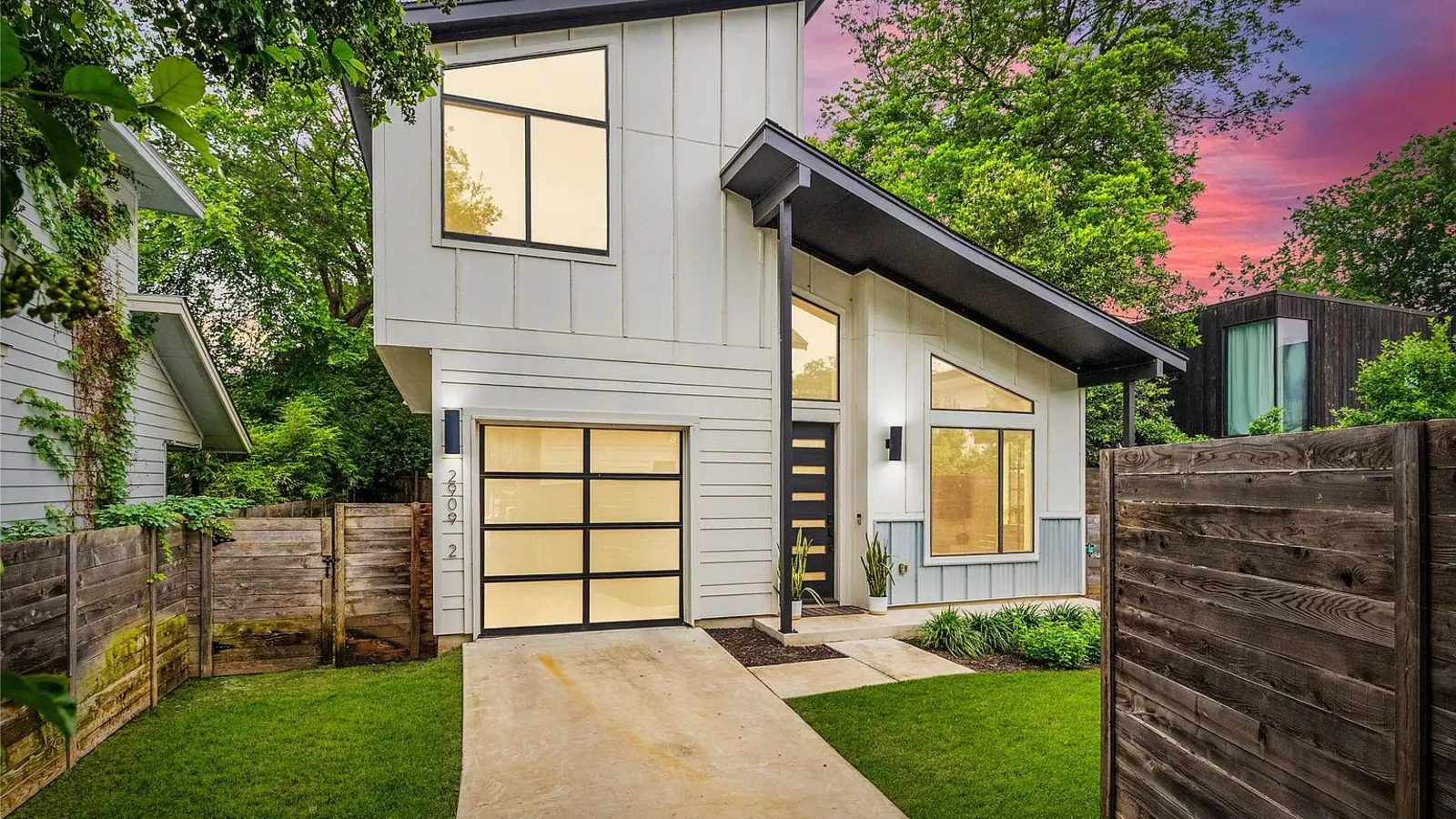 Modern white East Austin home with black trim, wood fencing, mature trees and a pink sunset sky.